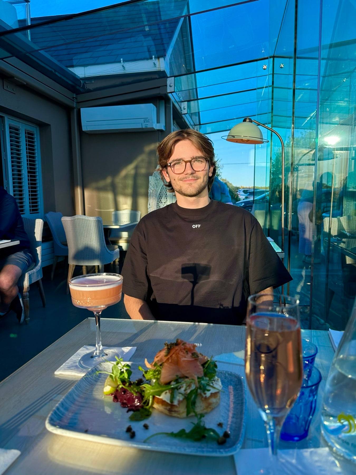 Sinclair smiling at a table by a glass wall, in a black T-shirt.